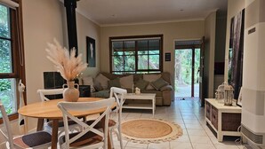 A cozy living room with a wooden dining table, white chairs, a beige sofa, and a large window with a view of greenery. - Brook Cottage@FernglenForestRetreat (Mount Dandenong)