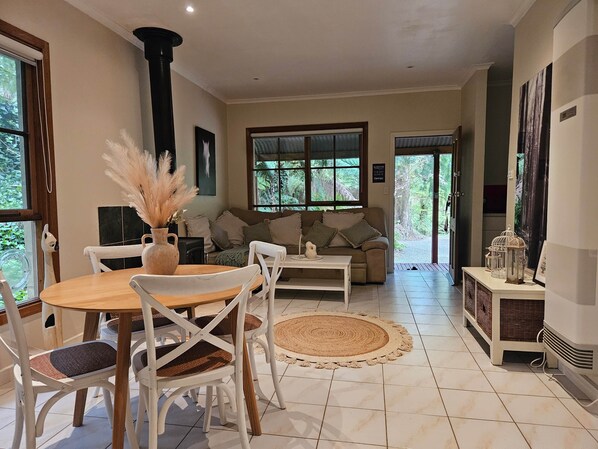 A cozy living room with a wooden dining table, white chairs, a beige sofa, and a large window with a view of greenery.