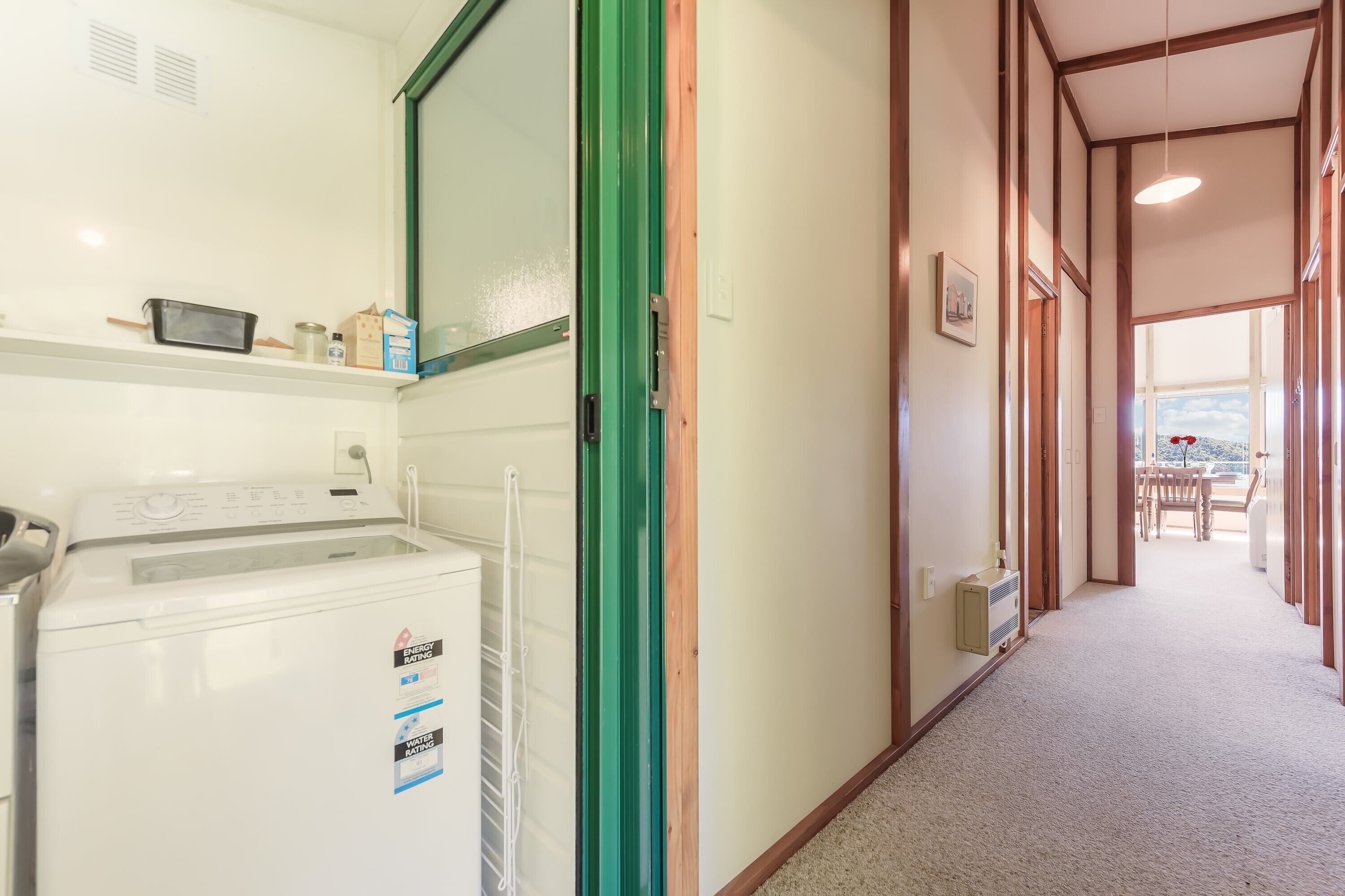 A laundry area with a washing machine, a microwave, and a shelf with items. A green door leads to a hallway with a view of a balcony.