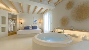 A modern bedroom with a large bathtub, a bed with blue bedding, and a walk-in closet with hanging robes. - Supreme Suite with Spa Bath (Naxos)