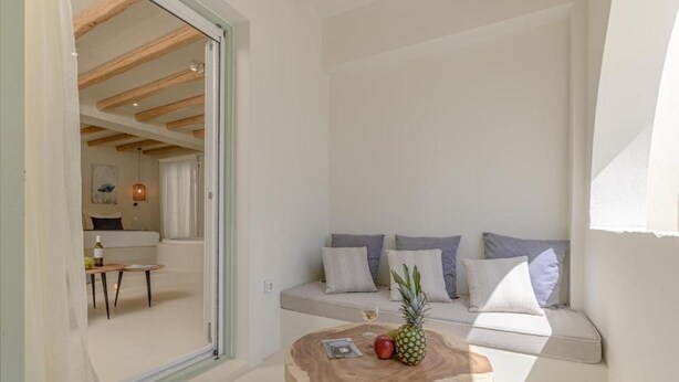 A modern living room with a wooden coffee table, a sofa with cushions, and a dining area visible through an open door. - Supreme Suite with Spa Bath (Naxos)