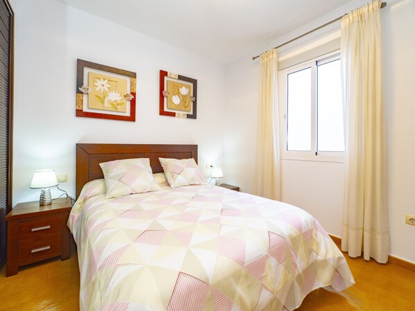 A neatly made bed with a geometric patterned bedspread, a wooden headboard, two bedside tables with lamps, and two framed floral artworks on the wall.