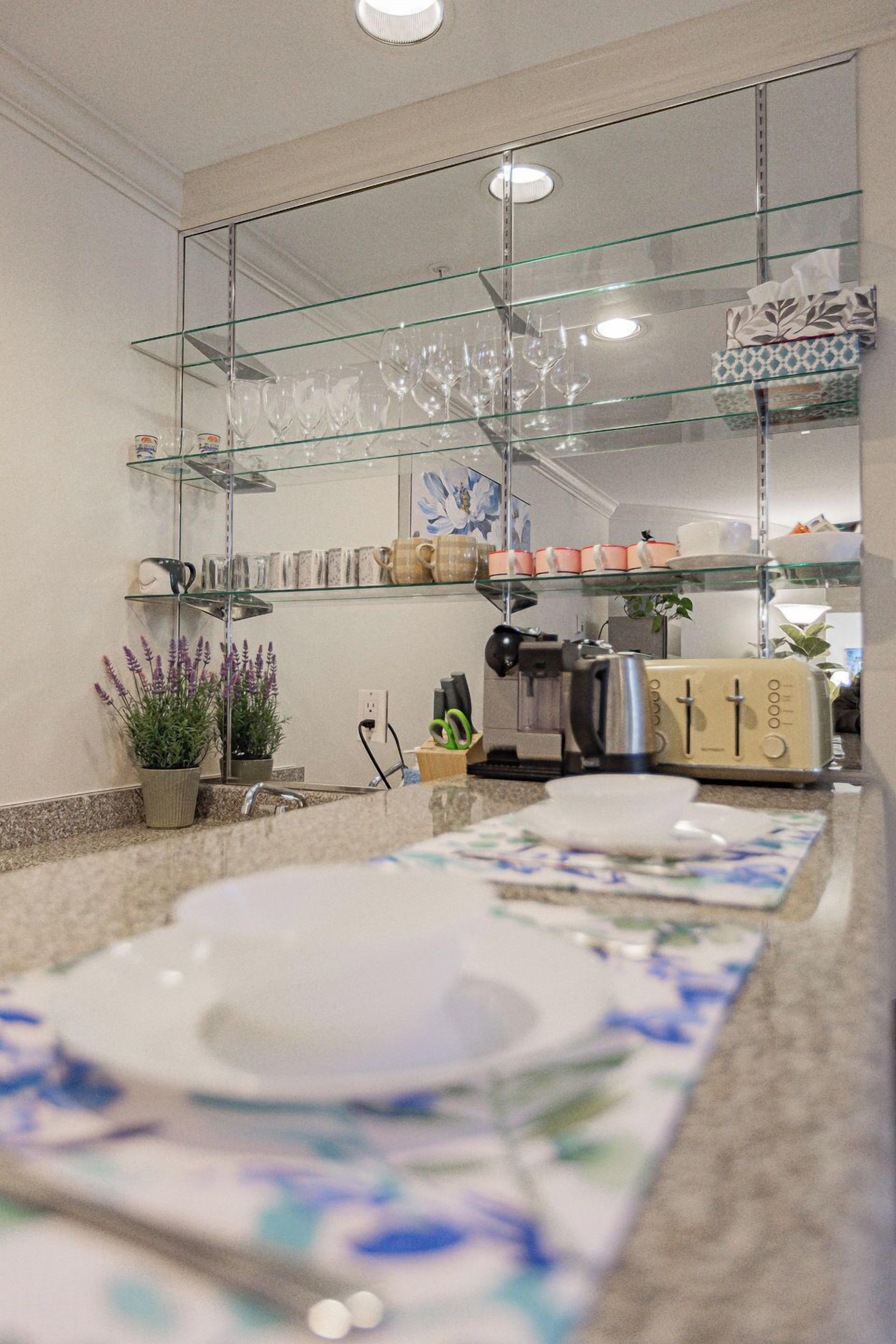 Ein Badezimmer mit einer Glasablage, die Gläser, eine Kaffeemaschine und einen Toaster beherbergt. Es gibt zwei Waschbecken mit Seifenschalen mit floralem Muster.