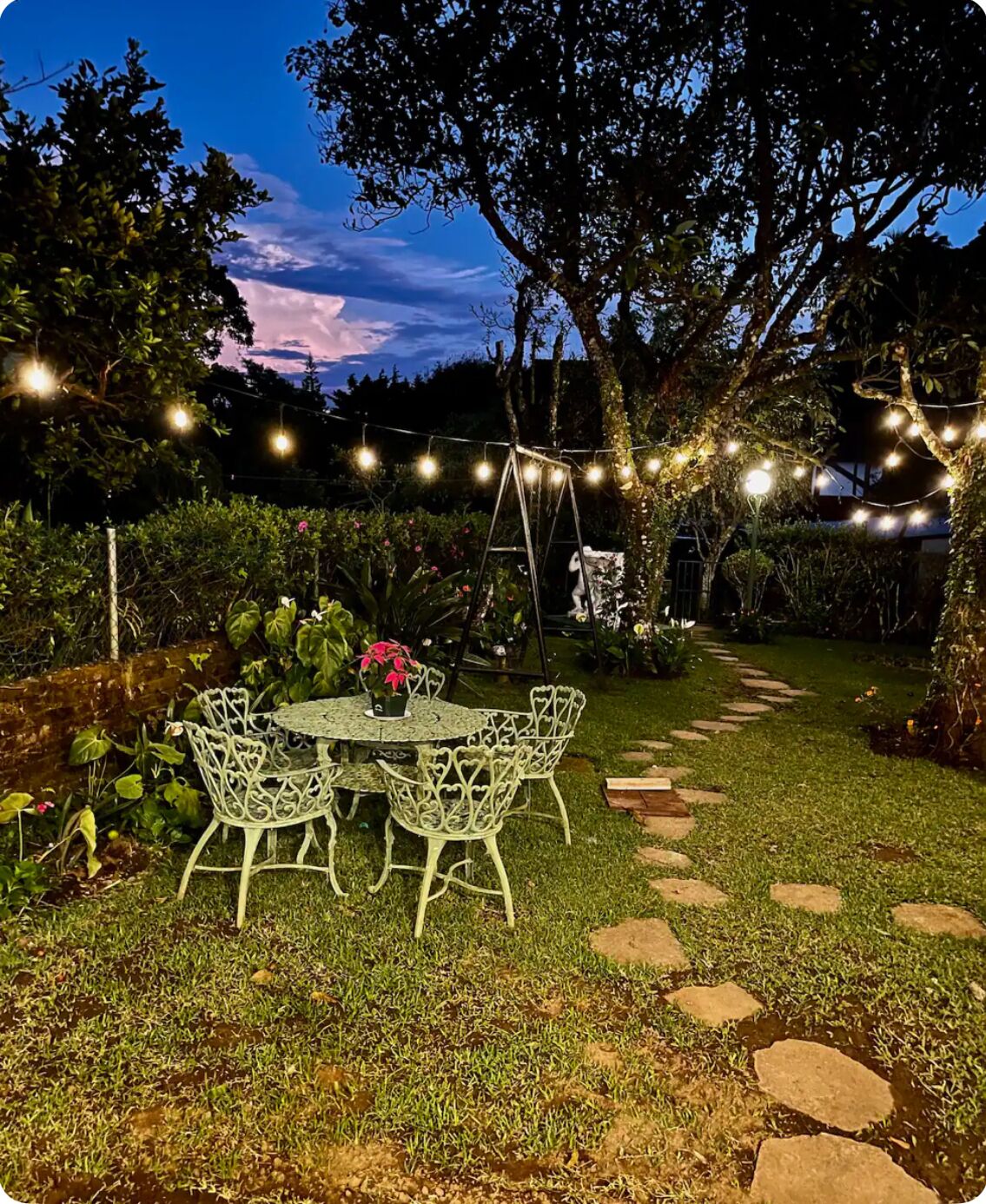 A garden patio with a round table and chairs, surrounded by greenery and stone paths.