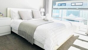 Een moderne slaapkamer met een groot bed, een nachtkastje, een lamp en uitzicht op een gebouw door het raam.