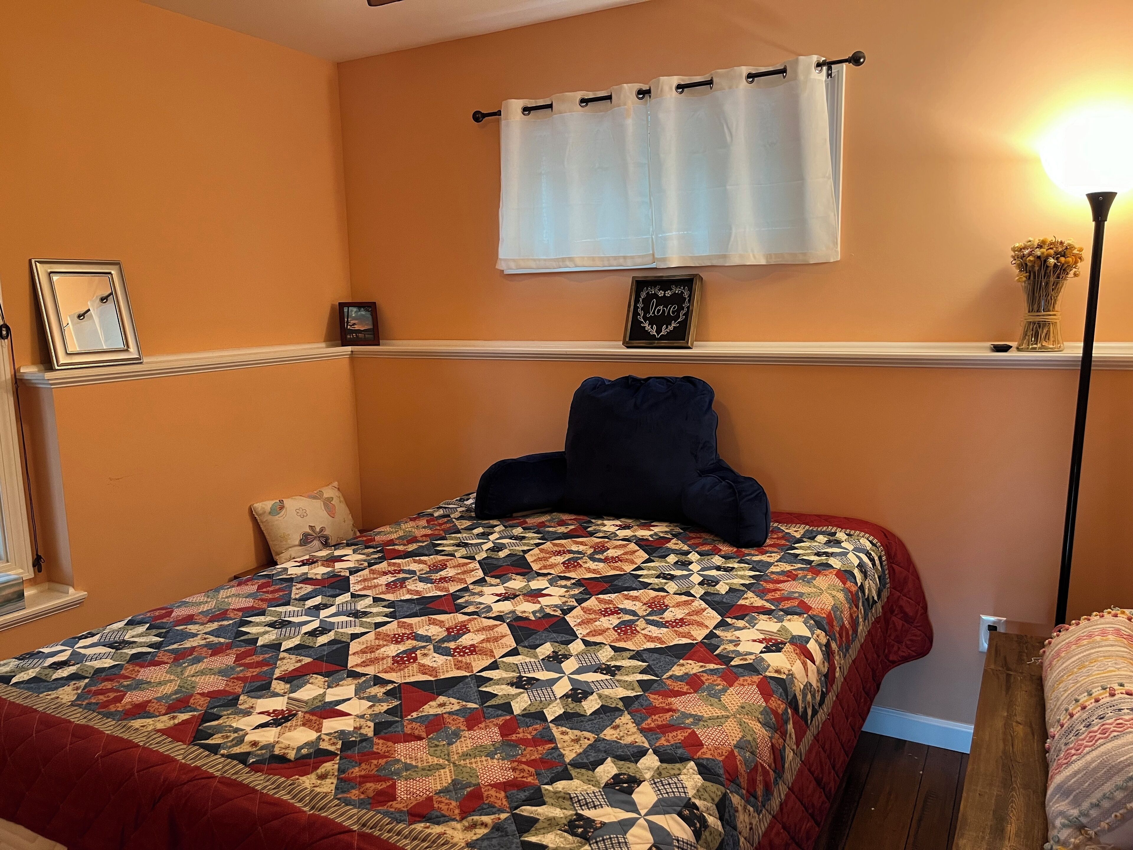 Une chambre avec un grand lit, une fenêtre ornée d’un rideau blanc, une lampe de chevet et une étagère fixée au mur, sur laquelle se trouve un vase.