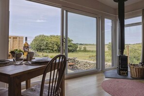 A dining area with a table set for two, a large window with a view of the outdoors, and a wood-burning stove. - The Hoot: A large, modern studio annexe on Anglesey (Llanfaelog)