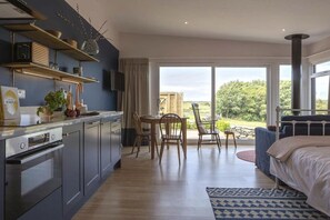 A modern bedroom with a large bed, a dining area with chairs, a kitchen with cabinets and a stove, and a view of the outdoors. - The Hoot: A large, modern studio annexe on Anglesey (Llanfaelog)