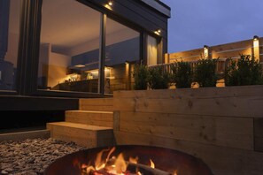 A modern outdoor seating area with a fire pit, wooden steps, and a view of a restaurant through large windows. - The Hoot : Quirky Pet-Friendly Studio Near Rhosneigr | Managed by Anglesey Holiday Lettings (Llanfaelog)