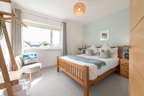 A bedroom with a wooden bed, a window with curtains, a chair, a bedside table with a lamp, and a wall-mounted picture.