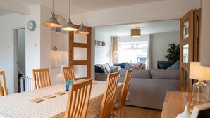 Un área de comedor con una mesa larga, sillas de madera e iluminación colgante. Se ve un sofá y una televisión al fondo.
