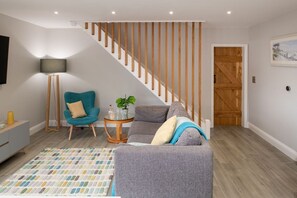 A living room with a grey sofa, a teal armchair, a small round table with a plant, and a staircase with wooden railings.