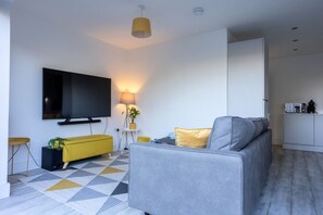 A modern living room with a sofa, a television, a lamp, and a kitchen area in the background.