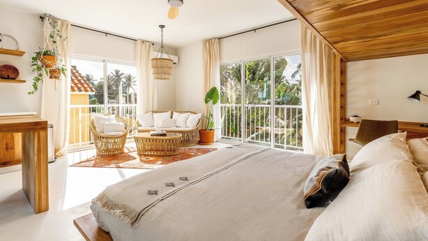 A bedroom with a large bed, a sitting area with wicker furniture, and a view of tropical vegetation through large windows. - Kite Beach Suite with Work Space and Full Kitchen (Cabarete)