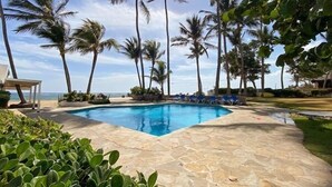 A swimming pool with a stone deck, surrounded by palm trees and a view of the ocean. - Kite Beach Suite with Work Space and Full Kitchen (Cabarete)