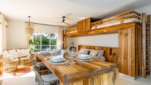A modern, open-plan living space with a wooden dining table set for a meal, a comfortable seating area with wicker furniture, and a view of lush greenery outside. - Kite Beach Suite with Work Space and Full Kitchen (Cabarete)