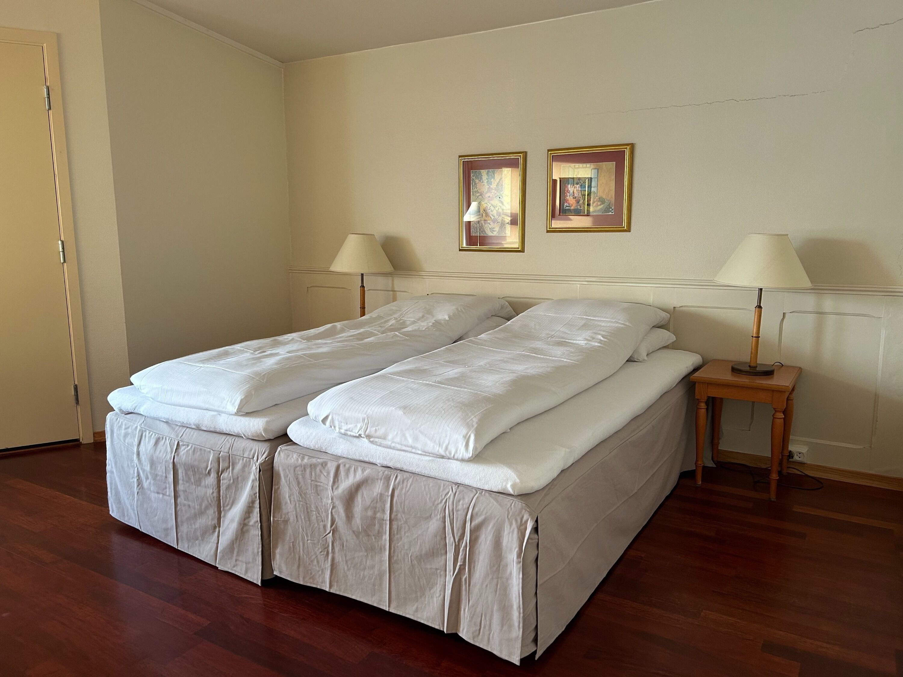 A double bed with a white duvet and matching skirt, two bedside lamps, and two framed artworks on the wall.