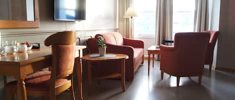 A hotel room with a TV mounted on the wall, a sofa, a chair, a small table with a lamp, and a potted plant.