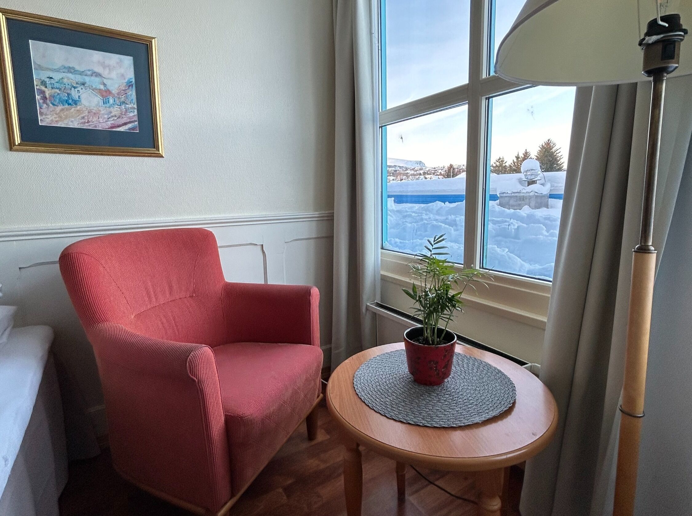 Um quarto com uma poltrona vermelha, uma pequena mesa de madeira com um vaso de planta, uma janela com vista para uma paisagem nevada e um quadro na parede.