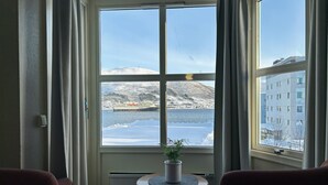 A room with a large window offering a view of a snowy landscape and a cruise ship. There is a small table with a plant in the center of the room. - Marina Hotel Sortland (Sortland)