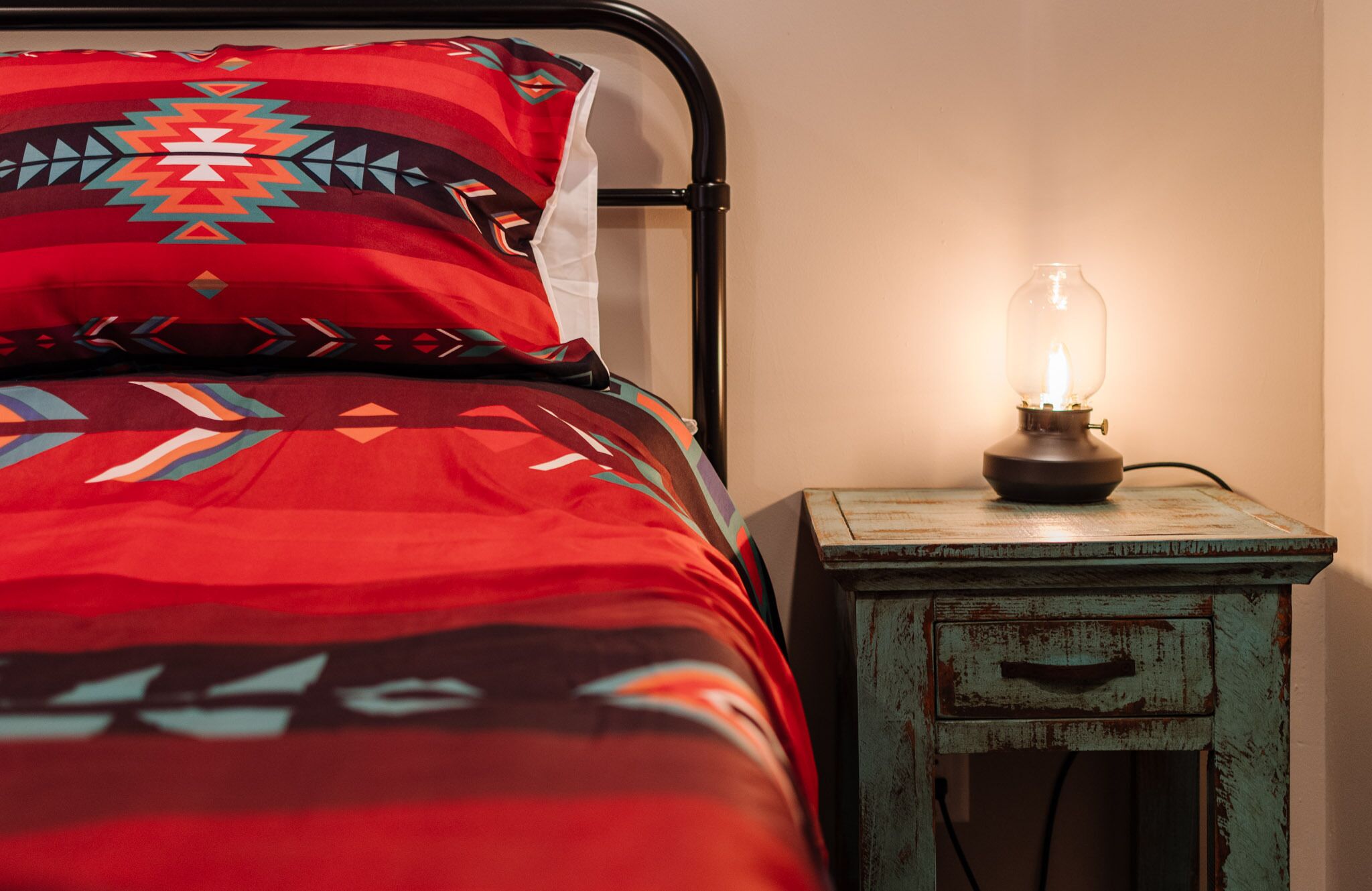 Una cama con una manta y almohadas estampadas, una lámpara de estilo antiguo sobre una mesita de noche de madera, y una pared con un diseño sencillo.