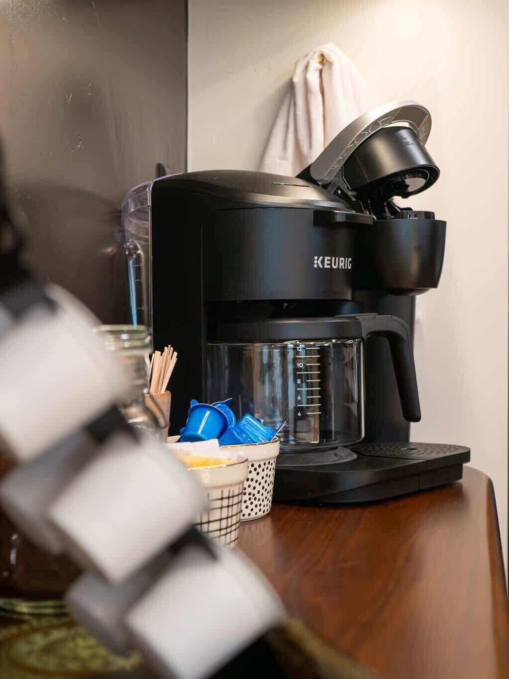 A Keurig coffee maker on a wooden table with a basket of reusable cups and a measuring cup.