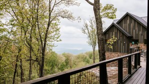 Une terrasse en bois avec une rambarde en métal, donnant sur une zone boisée et sur un bâtiment doté de fondations en pierre et d’un toit en métal.