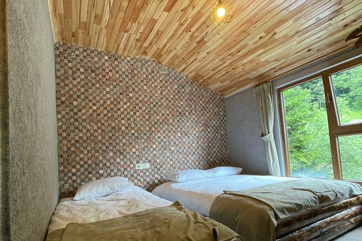 A room with a wooden ceiling, a mosaic headboard, two beds with white linens, and a large window with a view of greenery.