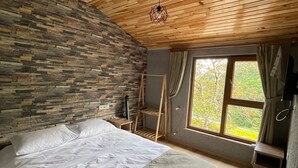 A bedroom with a stone wall, wooden ceiling, a bed, a window with curtains, and a small wooden shelf. - Bizim O'ra Küme Evleri & Bungalov (Maçka)