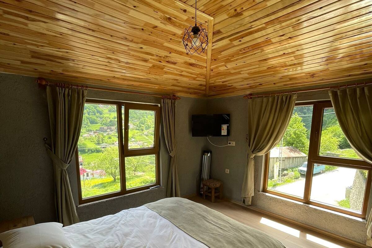 A bedroom with a wooden ceiling, a large bed, two windows with curtains, a flat-screen TV, and a small table.