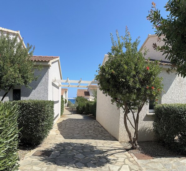 A stone-paved pathway leading to a white building with a tiled roof, surrounded by greenery and a clear blue sky.