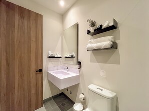 Ein modernes Badezimmer mit Holztür, weißem Waschbecken und einem Spiegel darüber. Handtücher sind auf Regalen zu sehen, außerdem eine Toilette.