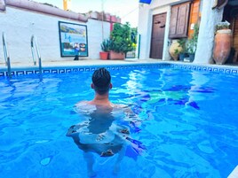 Un hombre nada en una piscina con borde de azulejos. Hay una escalera en el lado izquierdo de la piscina y plantas en macetas se ven al fondo.