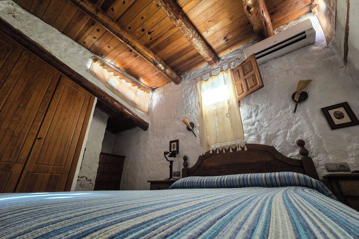A rustic bedroom with a wooden ceiling, a bed with striped bedding, a wooden wardrobe, and a window with a curtain.
