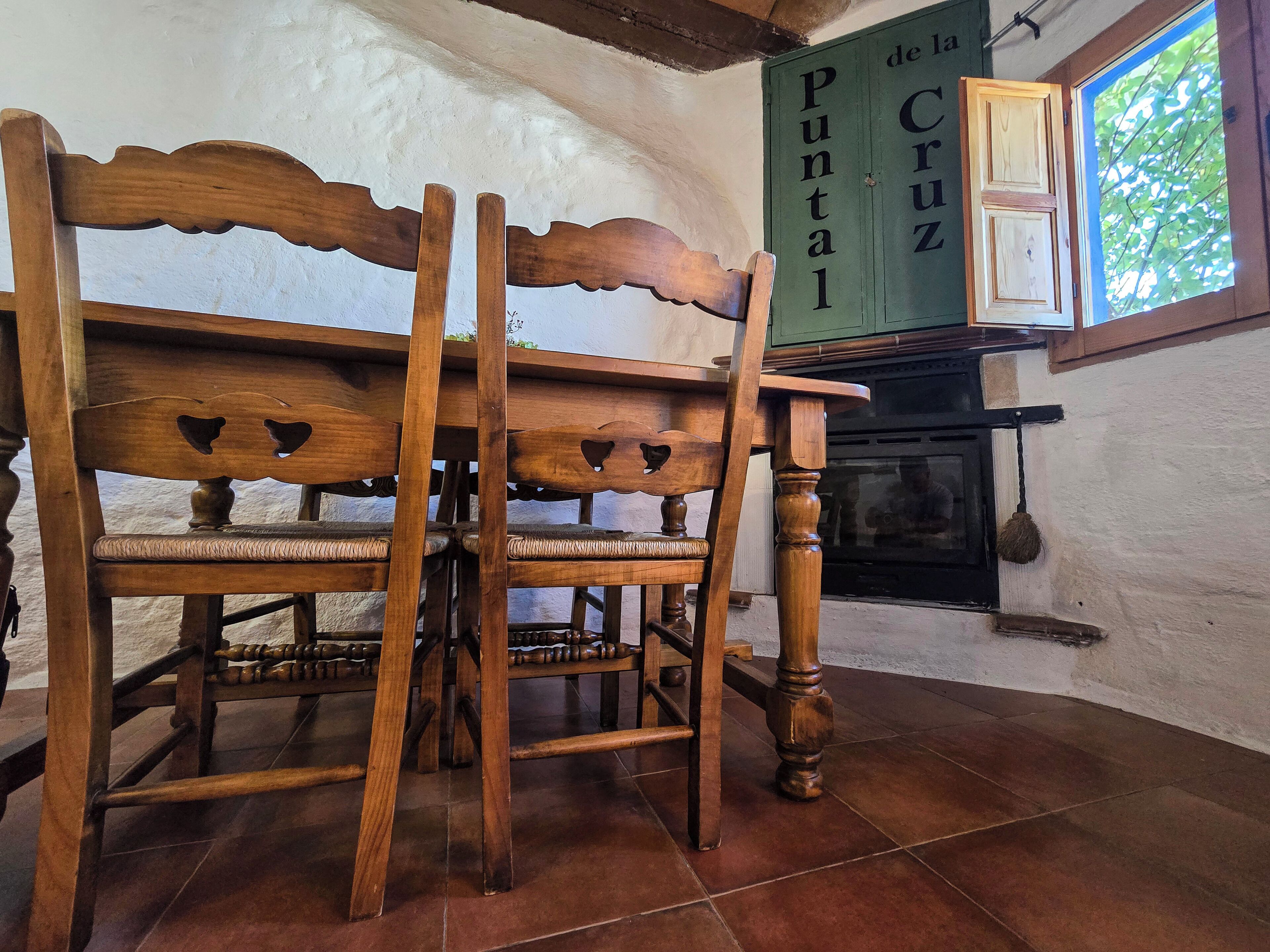 Un banc en bois, doté d’un dossier sculpté et d’une assise en osier, est placé dans une pièce dont le sol est recouvert de carreaux et où l’on peut voir une enseigne verte portant l’inscription « Puntal de Cruz ».