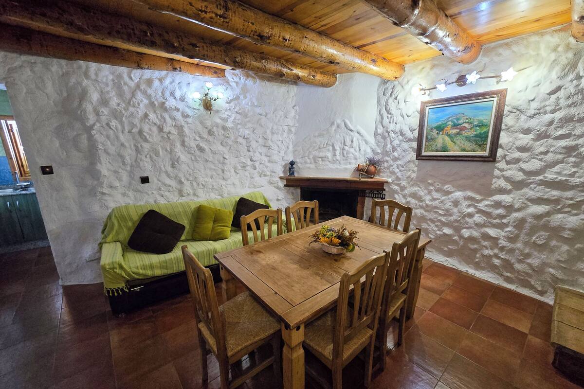 A rustic dining area with a wooden table and chairs, a green sofa with cushions, and a stone wall with a painting.