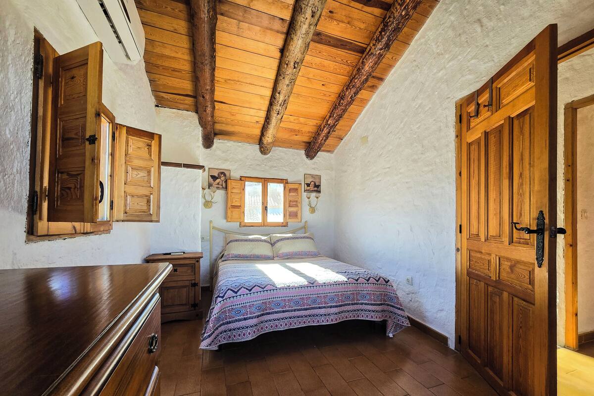 A bedroom with a wooden ceiling, a bed with patterned bedding, wooden doors, and a dresser.