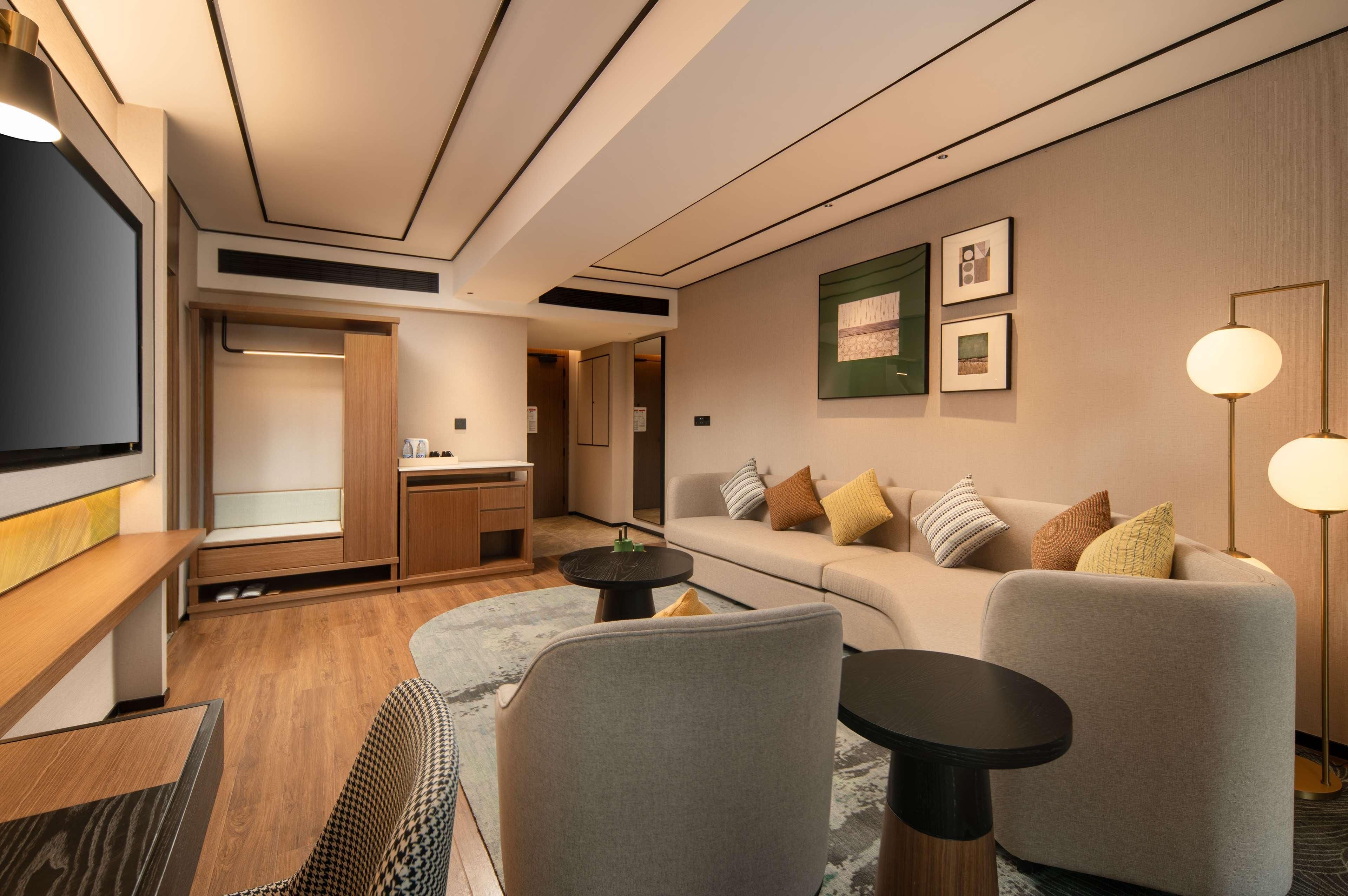 Quarto de hotel moderno com sofá, poltronas, mesa de centro, televisão e escrivaninha.