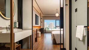 Una habitación de hotel moderna con una cama grande, un escritorio, una silla, un baño con lavabo y espejo, y vistas a la ciudad.
