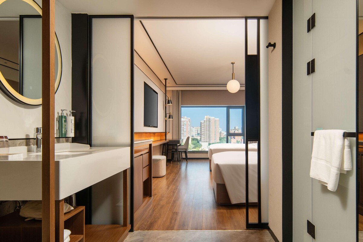 A modern hotel room with a large bed, a desk, a chair, a bathroom with a sink and mirror, and a view of the cityscape.