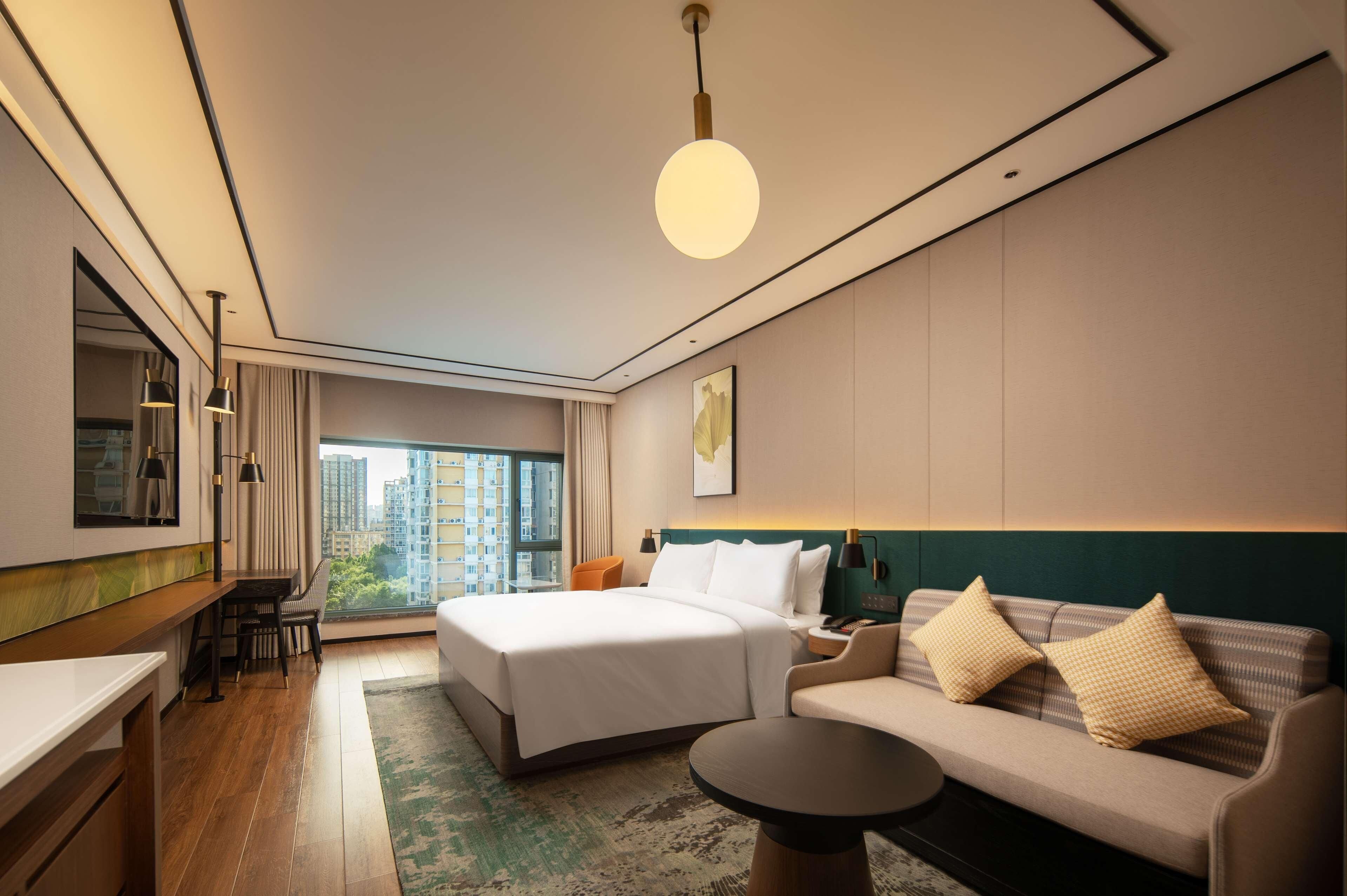 A modern hotel room with a large bed, a sofa, a desk, and a view of the city.