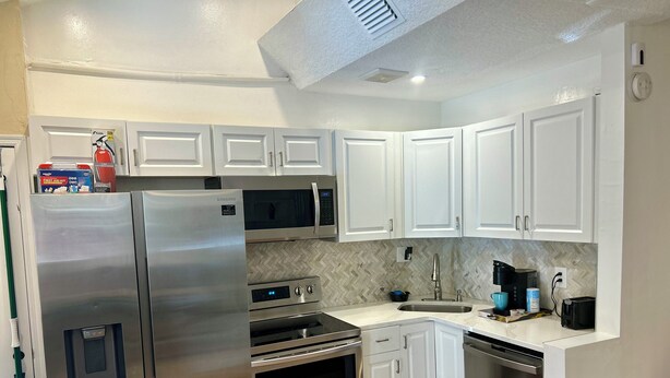 Una cocina moderna con armarios blancos, electrodomésticos de acero inoxidable, incluyendo un refrigerador, microondas, horno y lavavajillas, un salpicadero con motivo de espiga y suelo de baldosas.