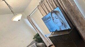 Una sala de estar con un sofá, un televisor mostrando la portada de un álbum musical, una lámpara en el techo y una ventana con cortinas.