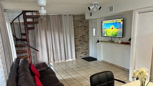 A living room with a brown leather sofa, a black chair, a dining table with a potted plant, a staircase, and a TV mounted on the wall displaying 'GOODNESS OF GOD'.
