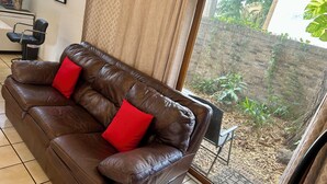 A brown leather sofa with red cushions, a tiled floor, a curtain, and a view of a garden through a glass door.