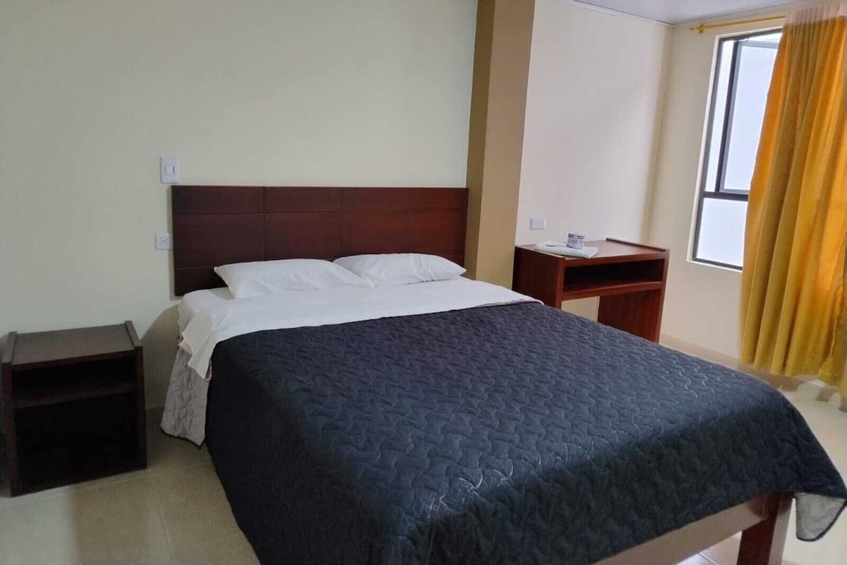 A hotel room with a large bed, a wooden headboard, a bedside table, and a window with curtains.