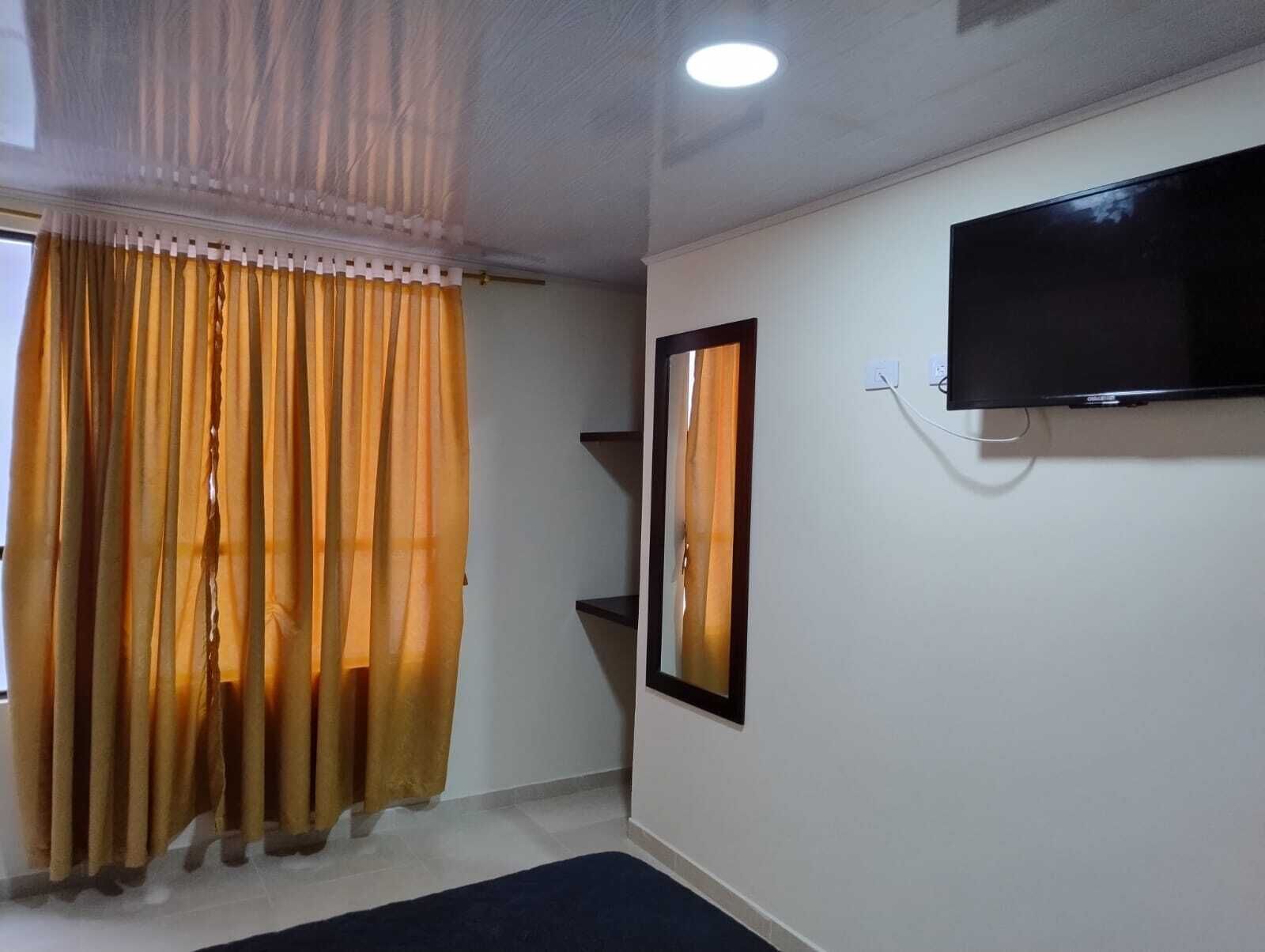 A room with a TV mounted on the wall, a window with curtains, and a small shelf.