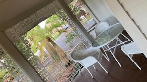 A covered outdoor seating area with white chairs and a table, surrounded by decorative metal railings and a view of a street with palm trees.