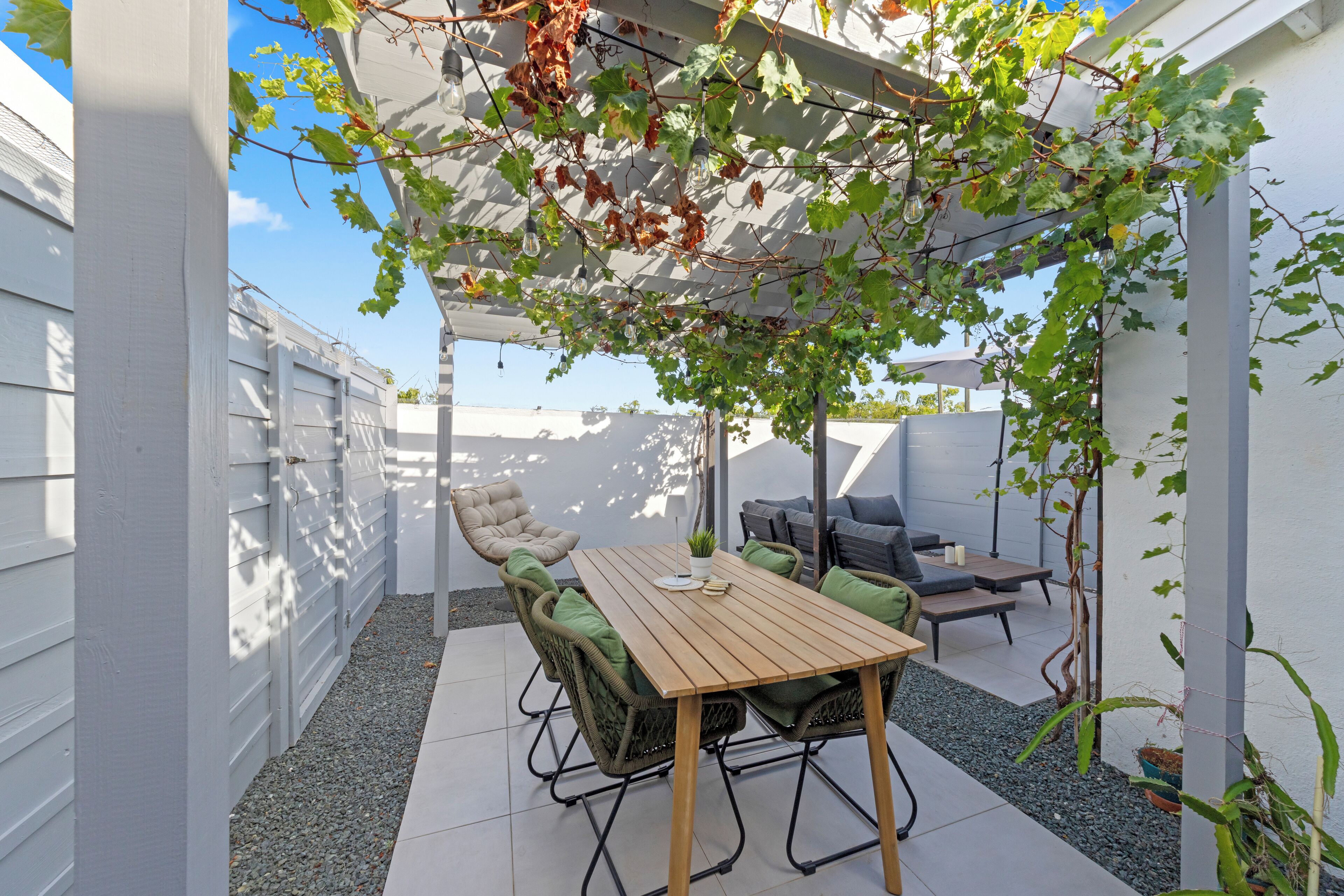 Een terras met een houten tafel, rieten stoelen en een pergola begroeid met klimop.