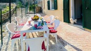 An outdoor patio with a table set for two, surrounded by white wicker chairs with red cushions, a large beige umbrella, and a bowl of fruit on the table. - Ca de Matette located just a few minutes' drive from beautiful Finalborgo. (Liguria)